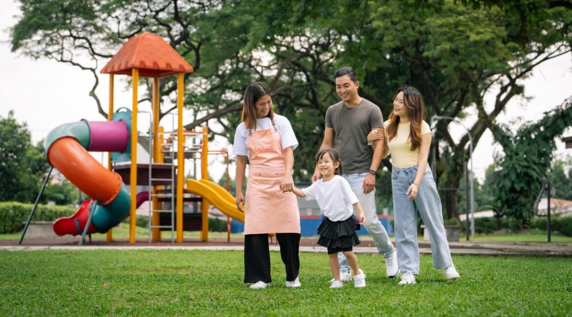 A smiling domestic helper from a top maid agency in Malaysia holding hands with a happy family in a playground located in Klang Valley home.