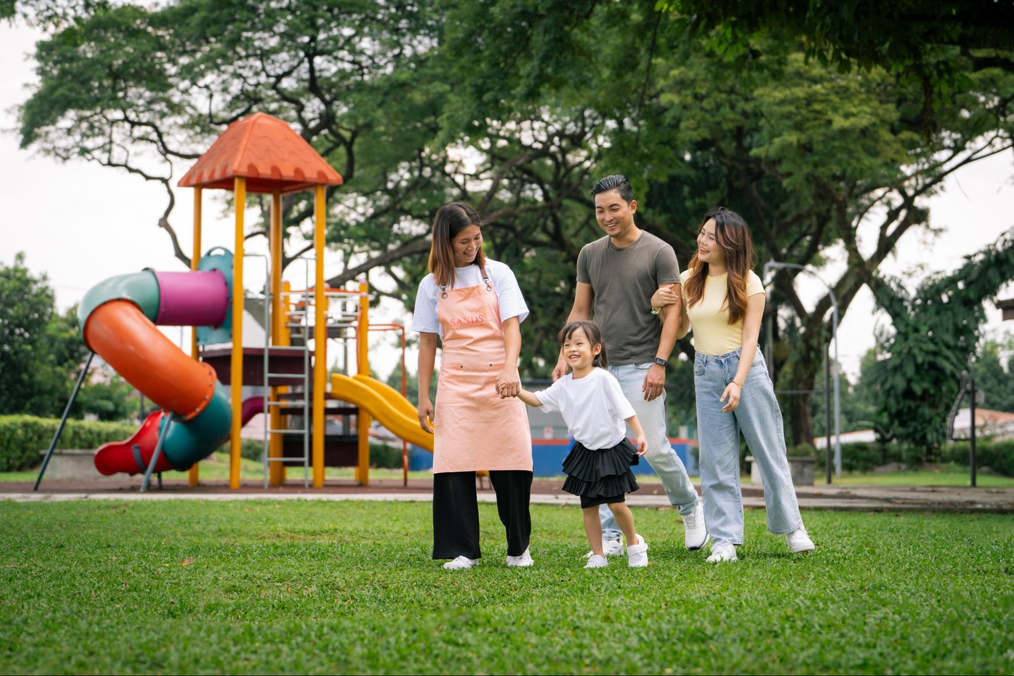 A smiling domestic helper from a top maid agency in Malaysia holding hands with a happy family in a playground located in Klang Valley home.